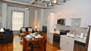 Uma cozinha moderna com eletrodomésticos de aço inoxidável, um espaço de refeições com mobiliário de madeira e uma sala de estar com sofá e luminária.