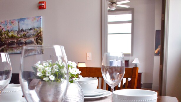 Uma mesa de jantar posta com pratos, tigelas e copos de vinho brancos, com um vaso de flores no centro.