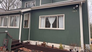 En grön byggnad med nummer 1323 på sidan, en veranda med trappsteg och ett fönster med en gardin.