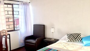 Ein Schlafzimmer mit einem Bett, einem Sessel, einem Nachttisch und einem Fenster mit Vorhängen.