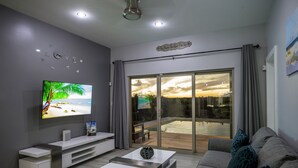 A modern living room with a flat-screen TV, a sofa, a coffee table, and a sliding glass door leading to a balcony. - SPECIAL DISC, 3BR PRIVATE VILLA, OCEAN VIEW, GREAT AMENITIES, STEPS TO THE BEACH (Long Bay Hills)
