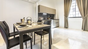 A modern dining area with a rectangular table set for four, flanked by black chairs. A kitchen with beige cabinets and a stainless steel range hood is visible in the background. - Elegant Fornasa Vecia Canal View R&R (Venecia)