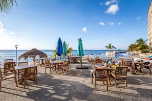 A beachfront patio with wooden tables and chairs, umbrellas, and a pool. - In The Heart of Cozumel (Cozumel)