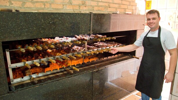 Una persona con delantal negro sosteniendo un pincho con carne sobre una parrilla abierta.