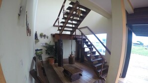 Una escalera de madera que conduce a un nivel superior, un área de descanso con sillas y un banco, y vistas del exterior a través de un amplio ventanal.
