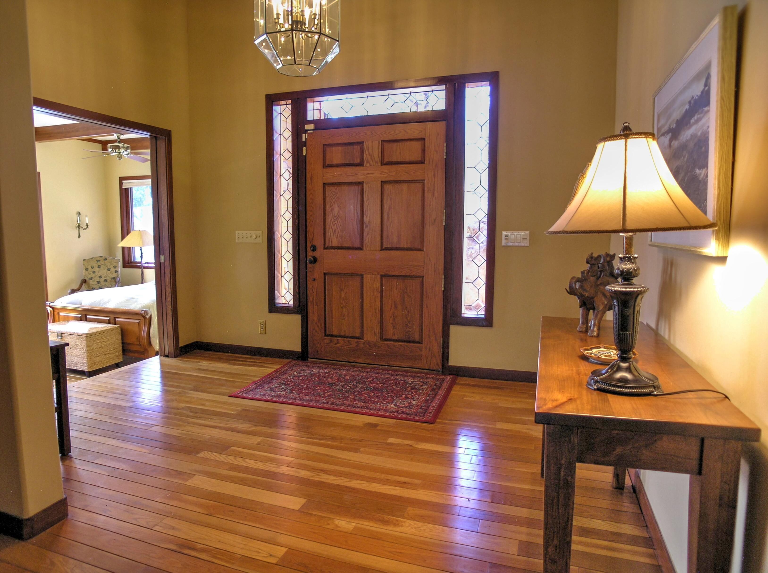 A wooden-floored hallway with a wooden door, a red rug, a lamp, and a small table with decorative items.