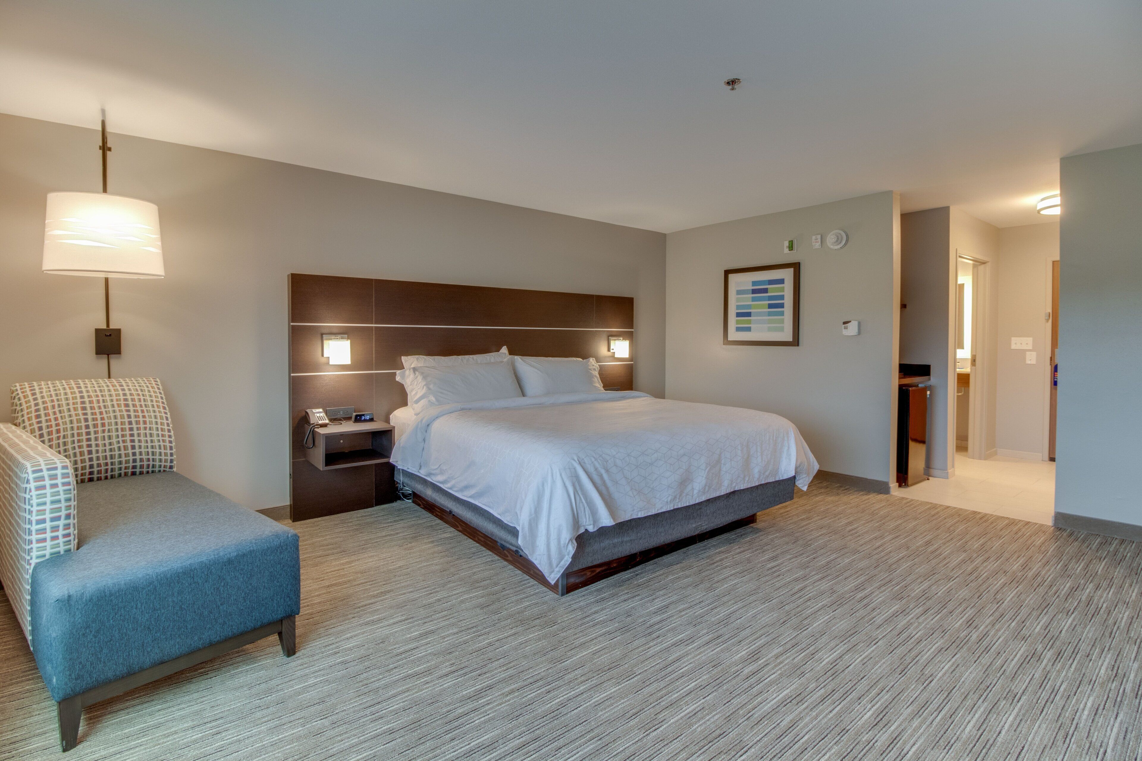 A hotel room with a bed, bedside table, lamp, chair, and a framed picture on the wall.