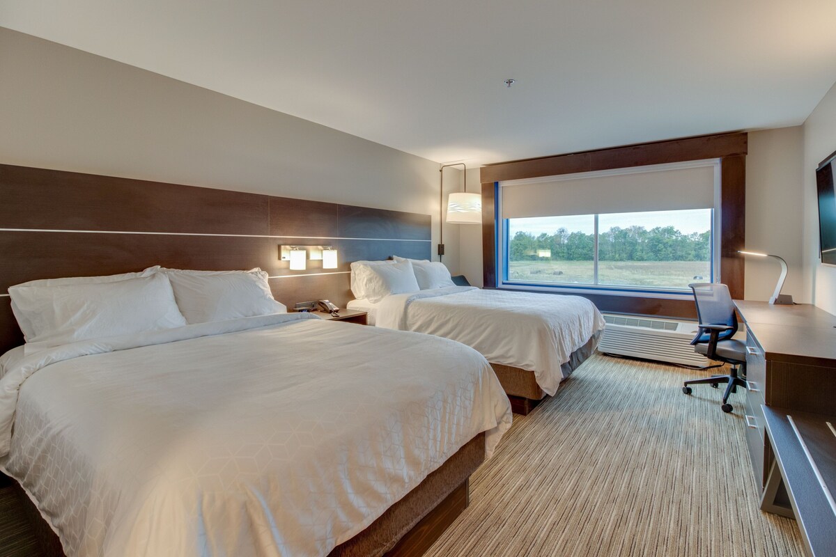 A hotel room with two beds, a desk, a chair, a large window with a view of the outdoors, and a flat-screen TV mounted on the wall.