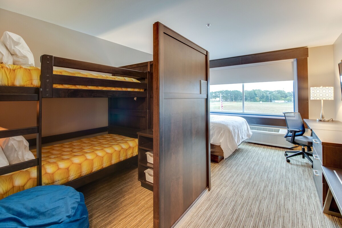 A hotel room with bunk beds, a desk, a chair, a lamp, and a large window with a view of the outdoors.