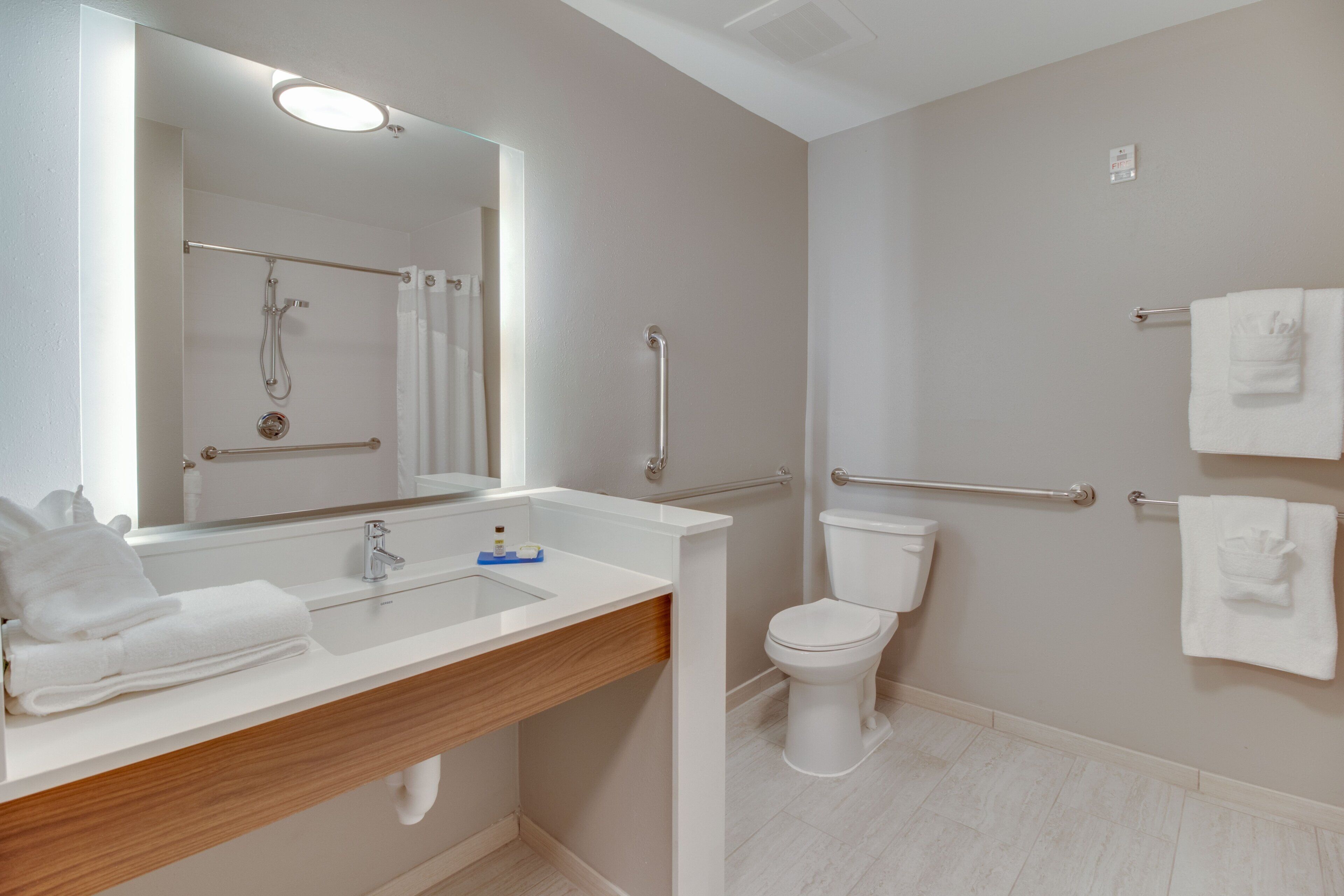Un baño moderno con lavamanos, espejo y retrete blancos. Hay dos toalleros con toallas, una zona de ducha con puerta de cristal y un pasamanos para accesibilidad.