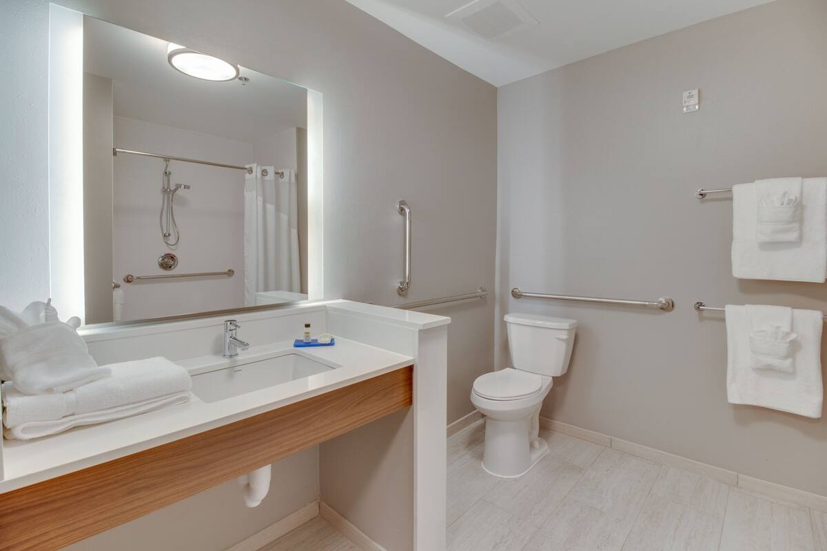 A modern bathroom with a white sink, mirror, and toilet. It includes a shower area with a grab bar and a towel rack.