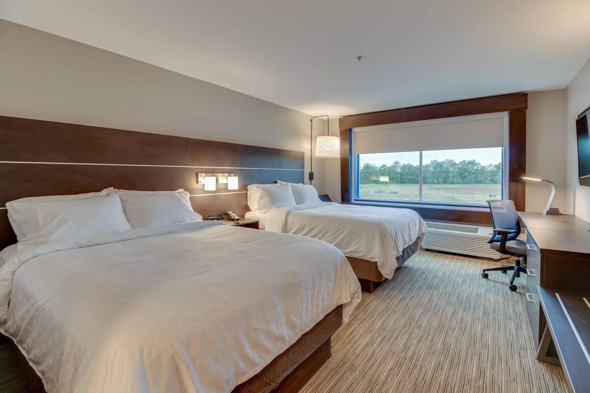 A hotel room with two beds, a desk, a chair, a large window with a view of the outdoors, and a flat-screen TV mounted on the wall.