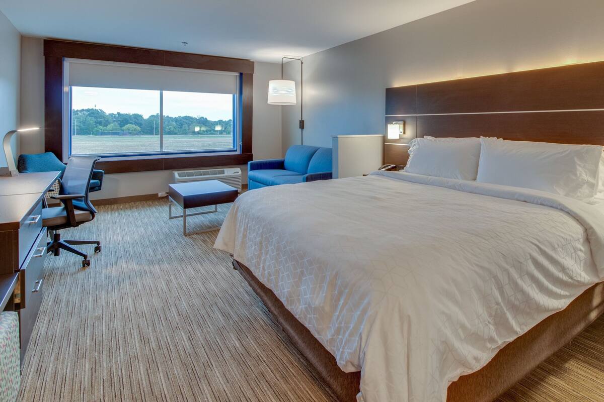 A modern hotel room with a large bed, a desk with a chair, a sofa, and a view of the outdoors through a large window.