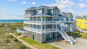 A multi-story beachfront house with balconies, a wooden deck, and a staircase leading to the upper levels. - Premier Oceanfront Home with Pool, Hot Tub, Elevator, Sauna, Game Room, Theater (Hatteras)