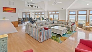 A spacious living room with a large striped sofa, a coffee table, and a red armchair. There are multiple windows offering a view of the ocean. - Premier Oceanfront Home with Pool, Hot Tub, Elevator, Sauna, Game Room, Theater (Hatteras)