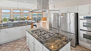A modern kitchen with a central island, stainless steel appliances, and large windows offering ocean views. - Premier Oceanfront Home with Pool, Hot Tub, Elevator, Sauna, Game Room, Theater (Hatteras)