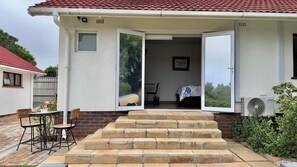 A house with a red-tiled roof, a set of stone steps leading to a door, a small table with chairs outside, and a window with a view of trees. - Somerset View Guest House (Cape Town)