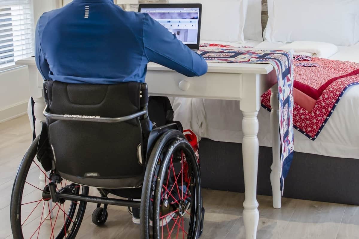 A person in a wheelchair at a white table with a laptop, a bed with a quilt, and an air conditioner on the wall.