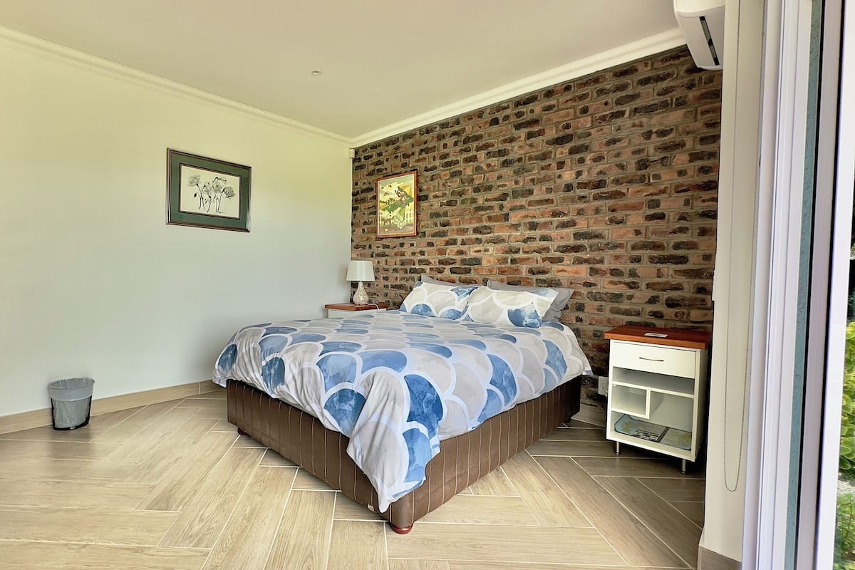 A bedroom with a brick wall, a bed with a patterned bedspread, a nightstand with a lamp, and a small white cabinet.