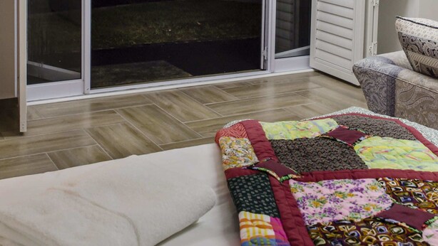 Una habitación con una puerta corrediza de vidrio que da a un espacio exterior, un edredón estampado en la cama y una pequeña cocina.