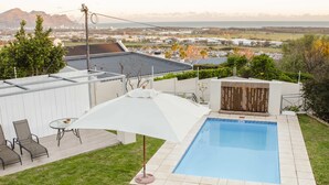 Patio trasero con piscina, terraza cubierta con sillas y vista a un paisaje urbano distante.