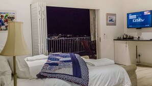 Habitación de hotel con cama, escritorio con televisor, balcón con barandilla y vista nocturna a la ciudad.