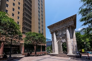 Una plaza pública con un monumento clásico, rodeada de árboles y edificios modernos.