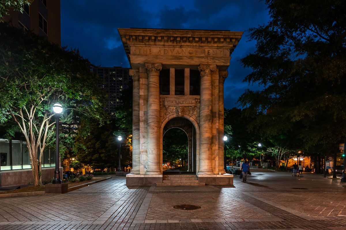 Un monument de marbre surmonté d’une arche, entouré d’arbres et d’éclairages publics, dans une place de la ville, de nuit.