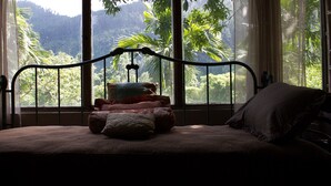 A bed with a metal frame, multiple pillows, and a view of a forest through large windows. - Lejartre's Retreat (Mount James)
