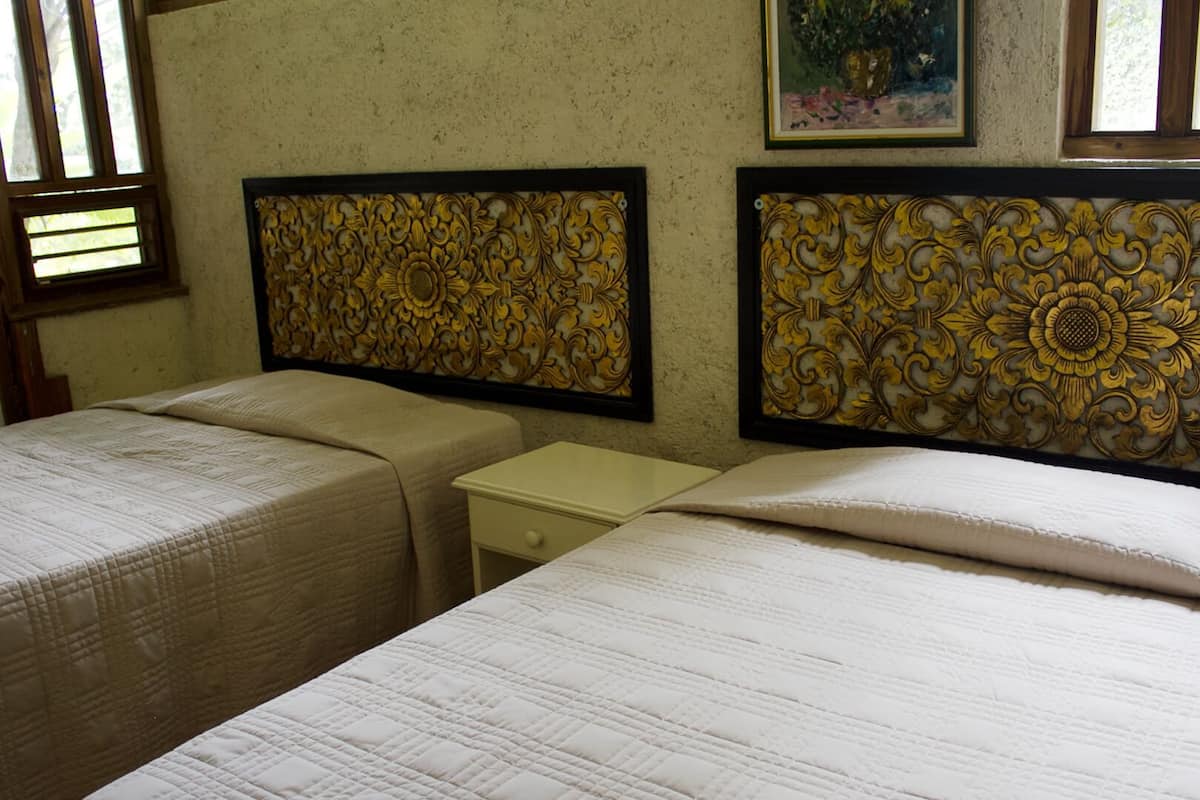 Ein Zimmer mit zwei Betten, jedes mit einem Kopfteil mit aufwendigen goldenen Verzierungen. Dazwischen steht ein kleiner Nachttisch, und links ist ein Fenster mit Jalousien zu sehen.
