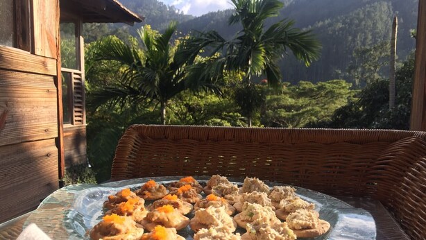 A wicker chair with a glass table displaying food items, set against a backdrop of a wooden structure and lush greenery. - Lejartre's Retreat (Mount James)