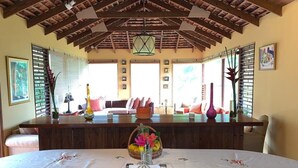 A dining area with a wooden ceiling, a central table with a floral tablecloth, and a view of the outside through large windows. - Lejartre's Retreat (Mount James)