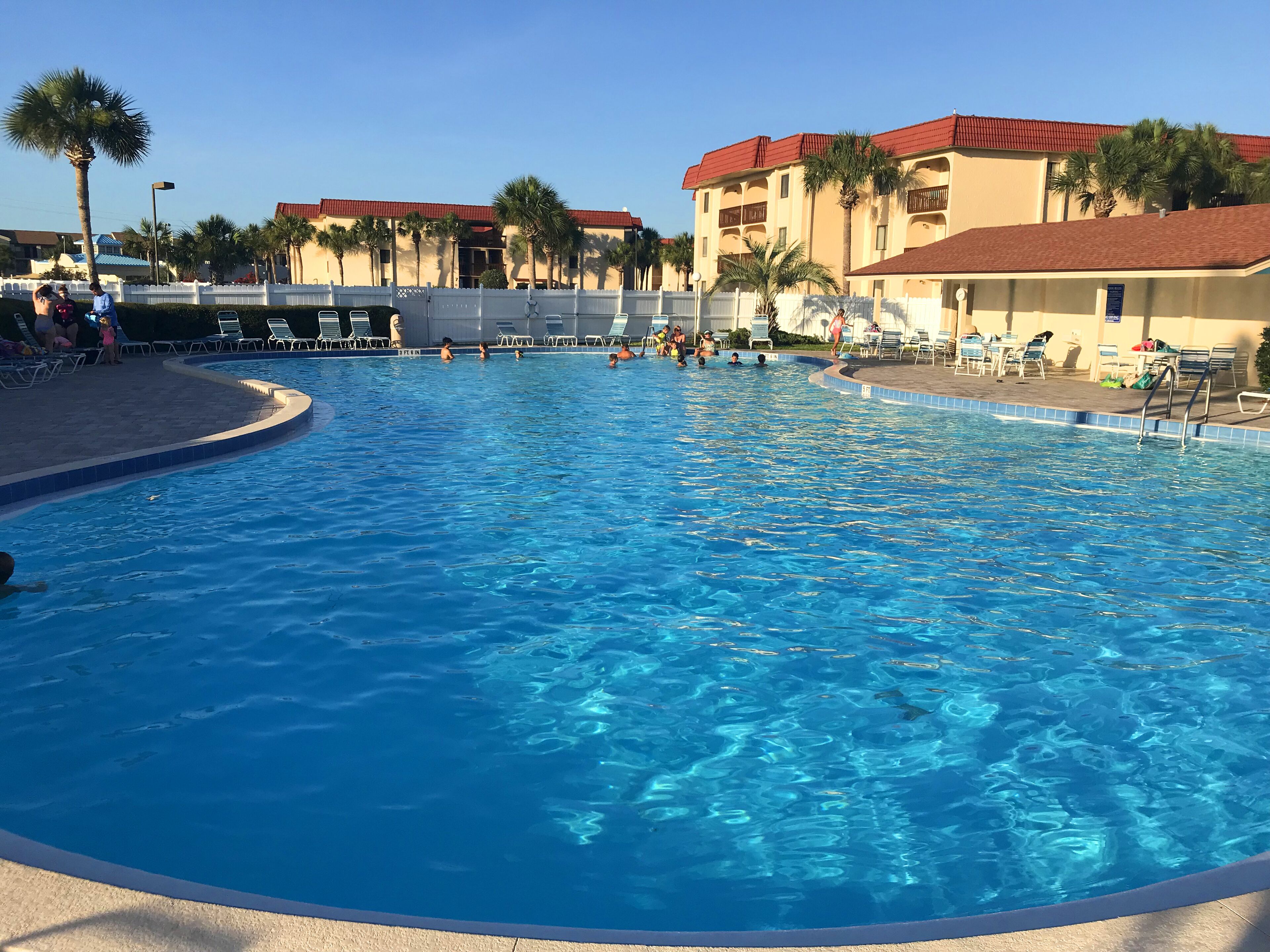 Una piscina con agua azul cristalina, rodeada de palmeras y un edificio con techo rojo.