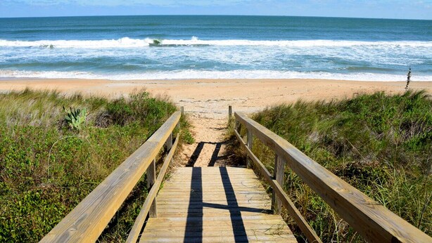 Un paseo marítimo de madera que conduce a una playa de arena, con olas del océano al fondo.