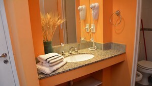 Een badkamer met een granieten aanrecht, een witte wastafel en een spiegel. Twee haardroger zitten aan de muur, een handdoekrek en een toilet zijn zichtbaar.