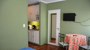 Een compacte kamer met een kitchenette, een eettafel met een tafelkleed met patroon, en een aan de muur bevestigde televisie.