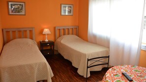 A room with two single beds, a small table with a colorful tablecloth, and a lamp on a nightstand.