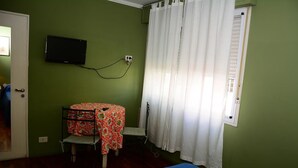 Een kamer met groene wanden, een bed met een rood-wit gebeddeken, een aan de muur bevestigde televisie en een raam met witte gordijnen.
