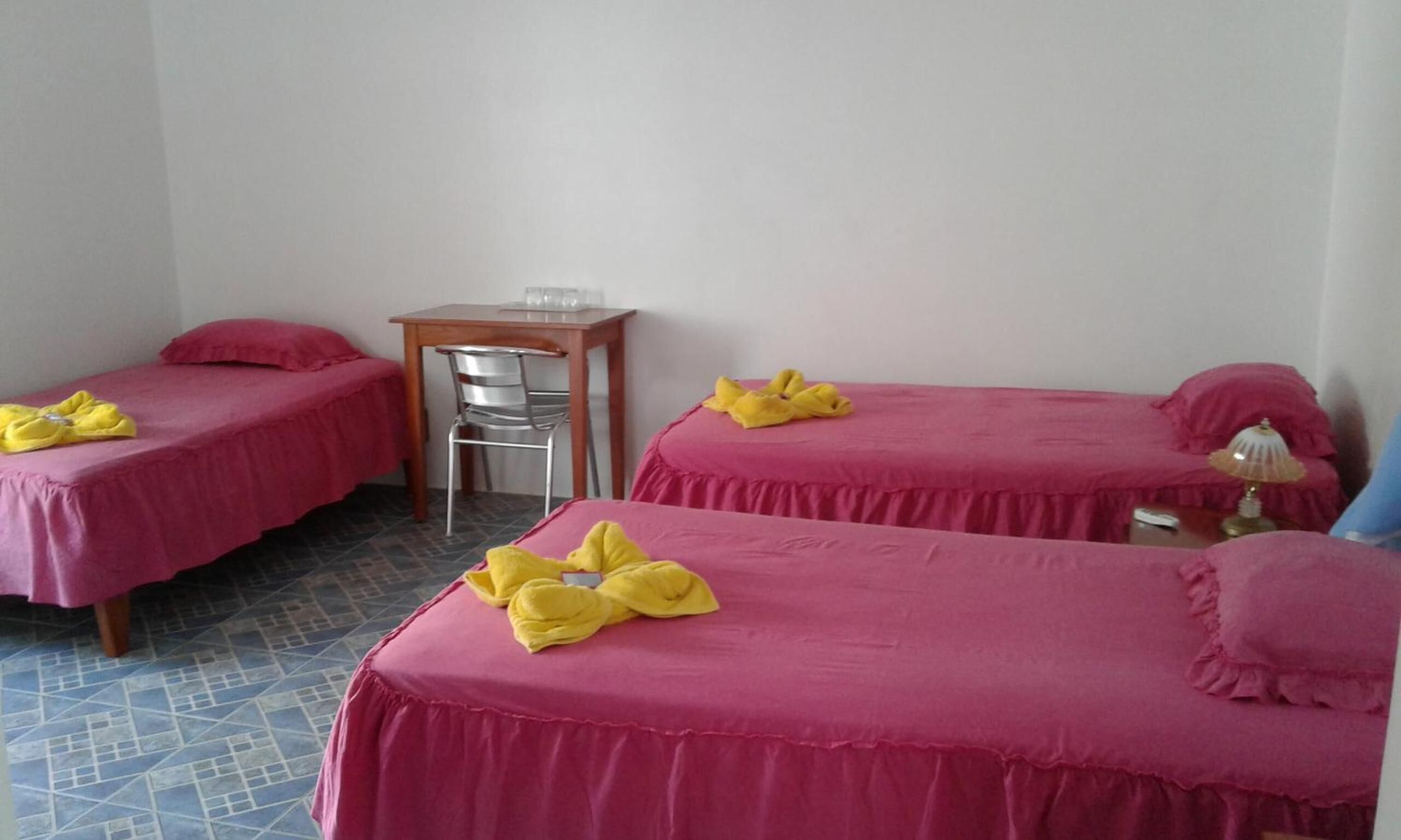 Ein Hotelzimmer mit zwei Einzelbetten, beide mit rosa Bettdecken und einem gelben Handtuch. Es gibt einen Holztisch mit einem Stuhl, eine Lampe auf einem Nachttisch und einen Fliesenboden.