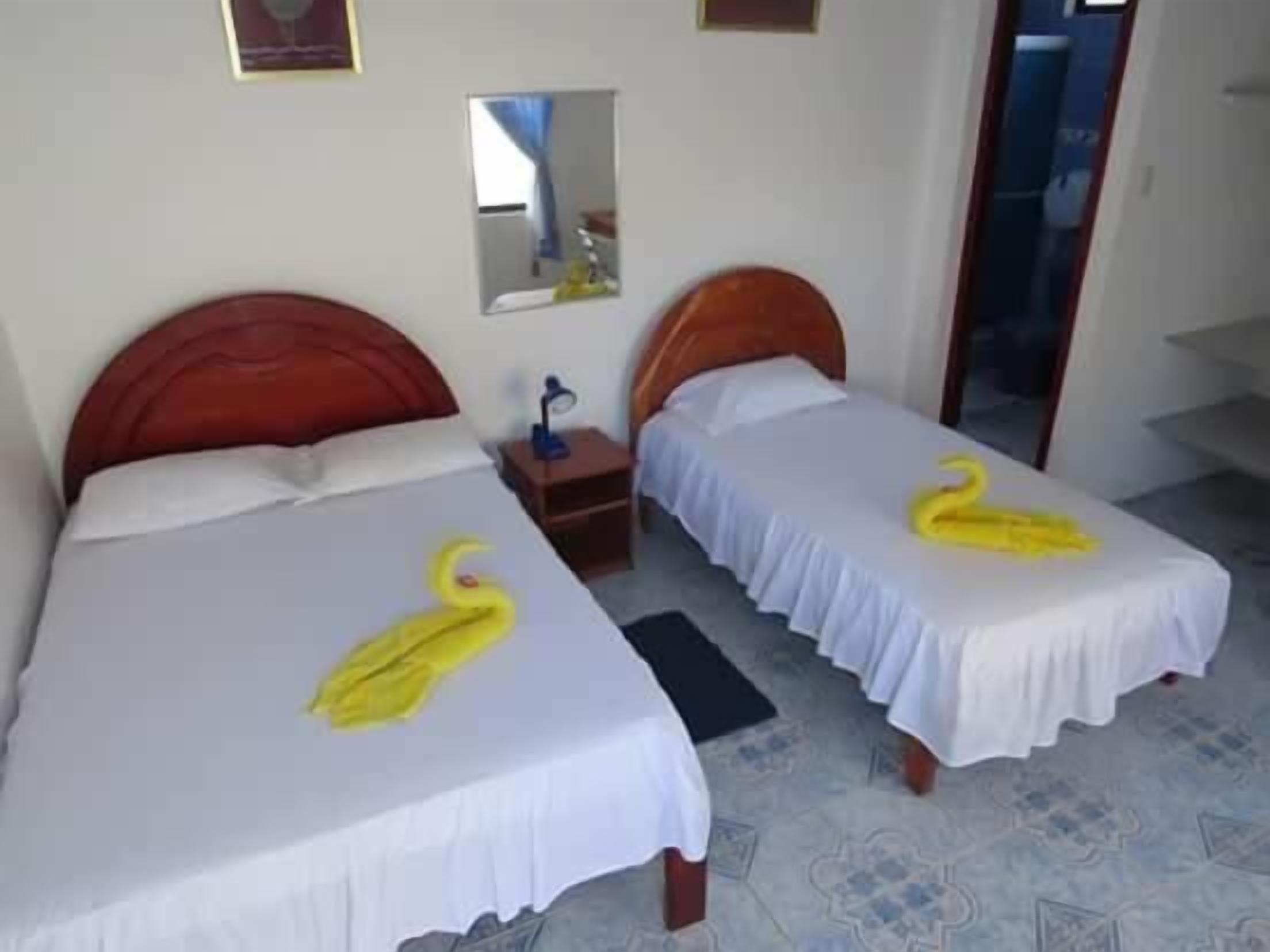 A hotel room with two beds, each with a white bedspread and a yellow towel shaped like a swan. There is a small bedside table with a lamp, a mirror on the wall, and a door leading to another room.