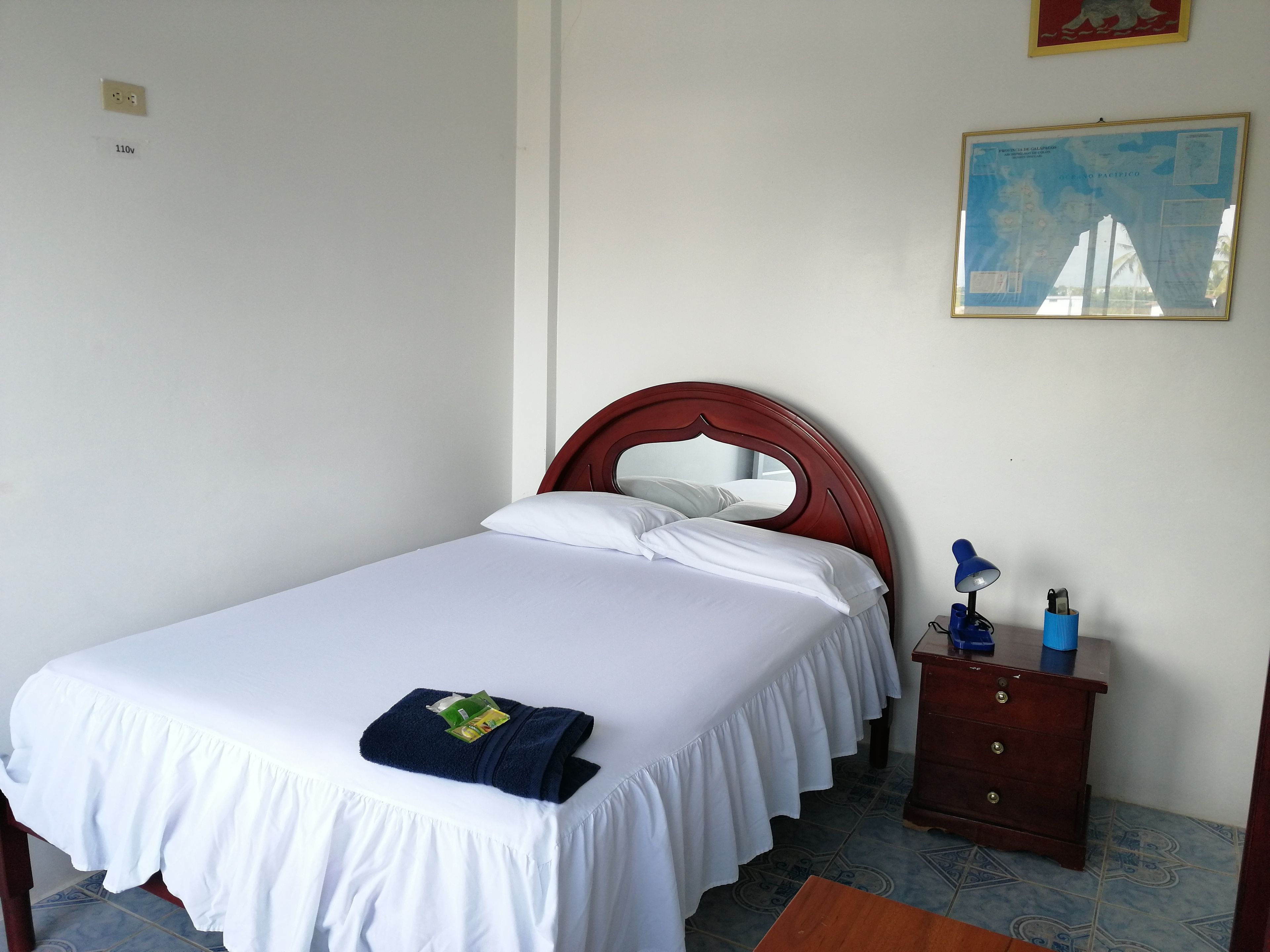 Una cama bien hecha con ropa de cama blanca y un cabecero rojo, un tocador de madera con una lámpara azul, y un mapa enmarcado en la pared.