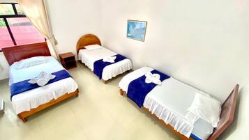 A hotel room with three beds, each with a white and blue bedspread and a white towel. There is a small wooden nightstand between the beds, and a window with a curtain.