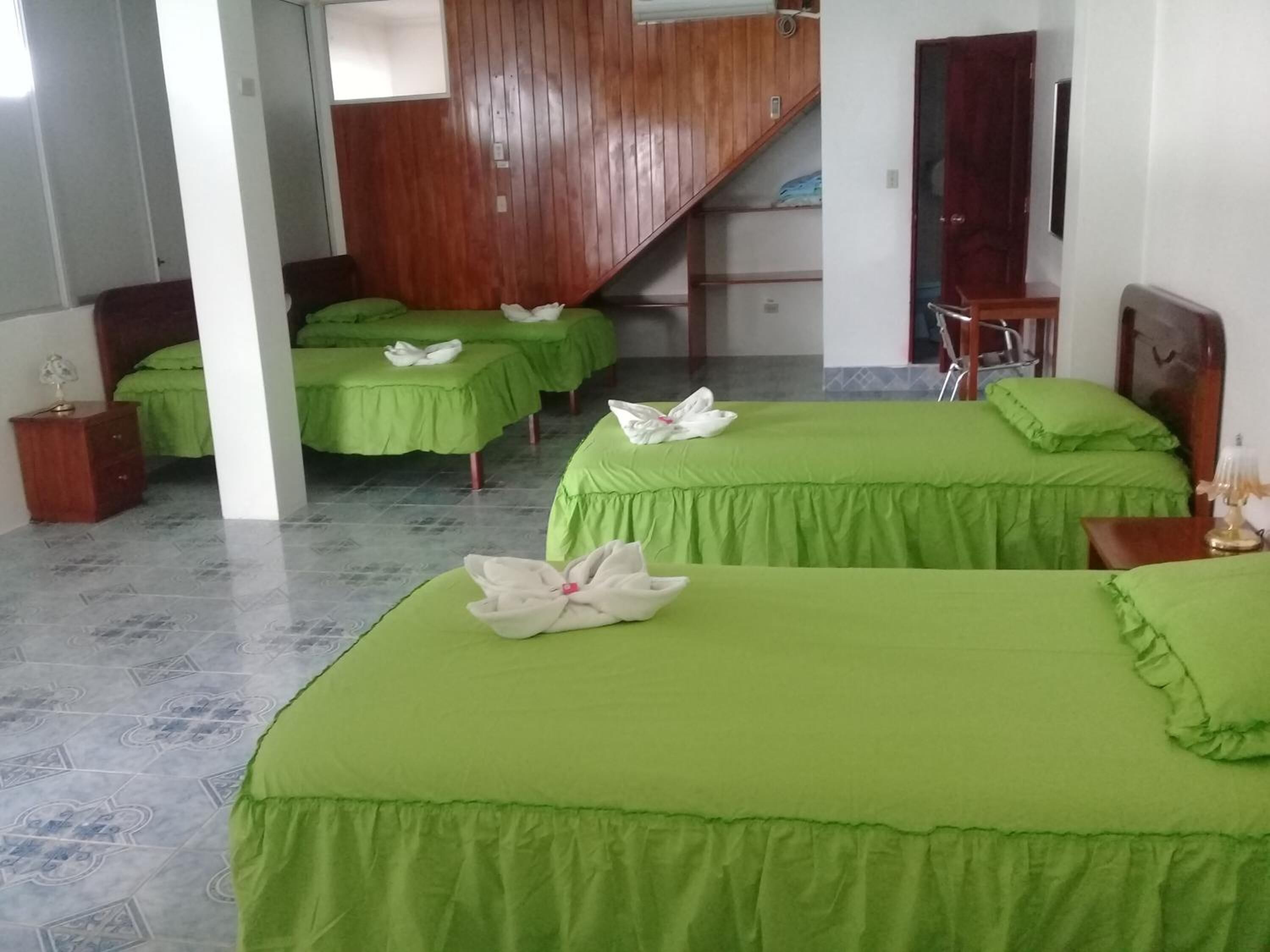 A hotel room with two beds, each with a green bedspread and white towels. There is a wooden staircase in the background, and a small wooden table with a lamp on the right side of the room.