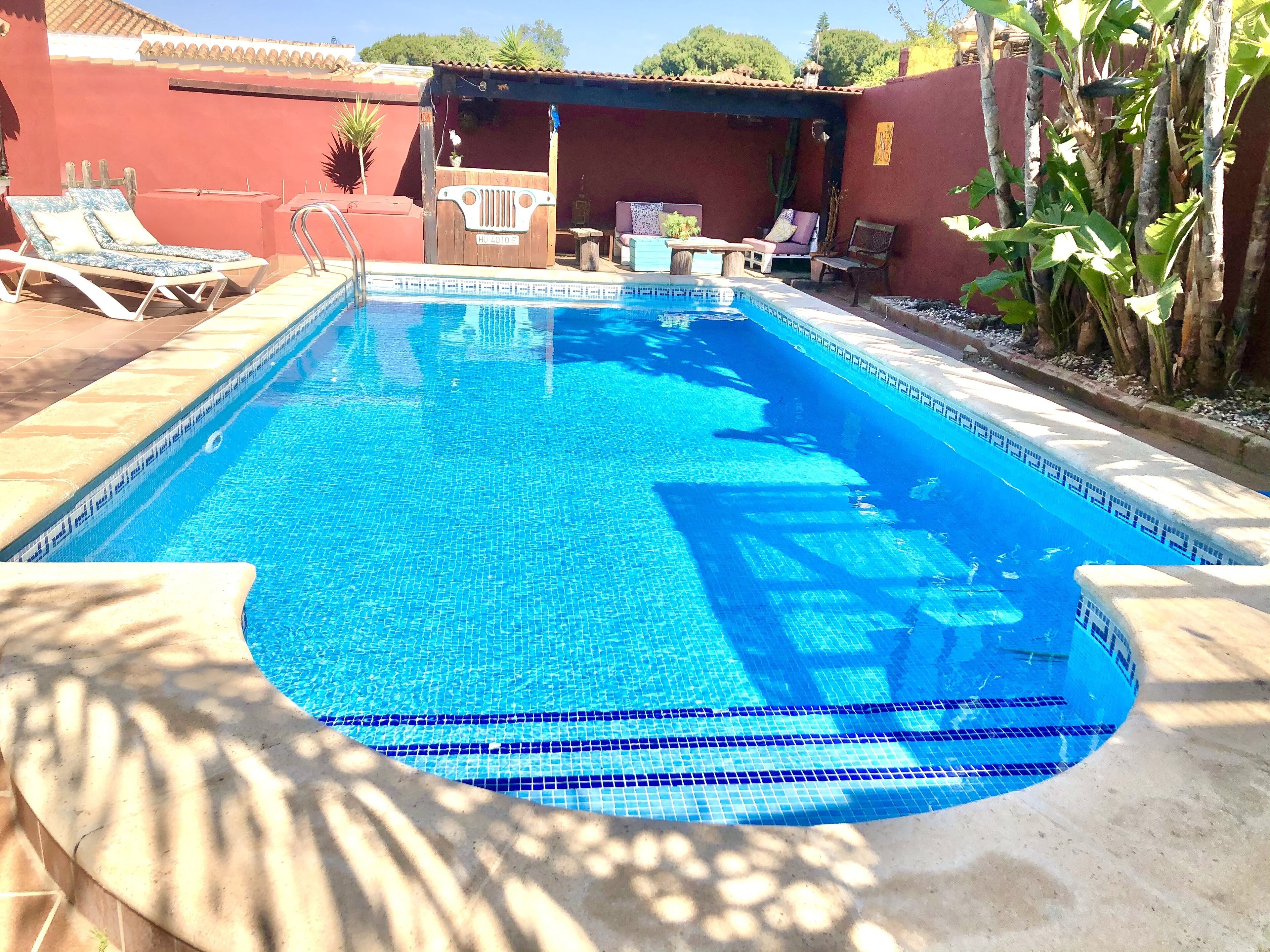 A rectangular swimming pool with a tiled edge, surrounded by lounge chairs and a covered seating area.