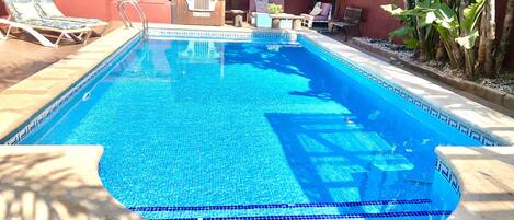 A rectangular swimming pool with a tiled edge, surrounded by lounge chairs and a covered seating area.