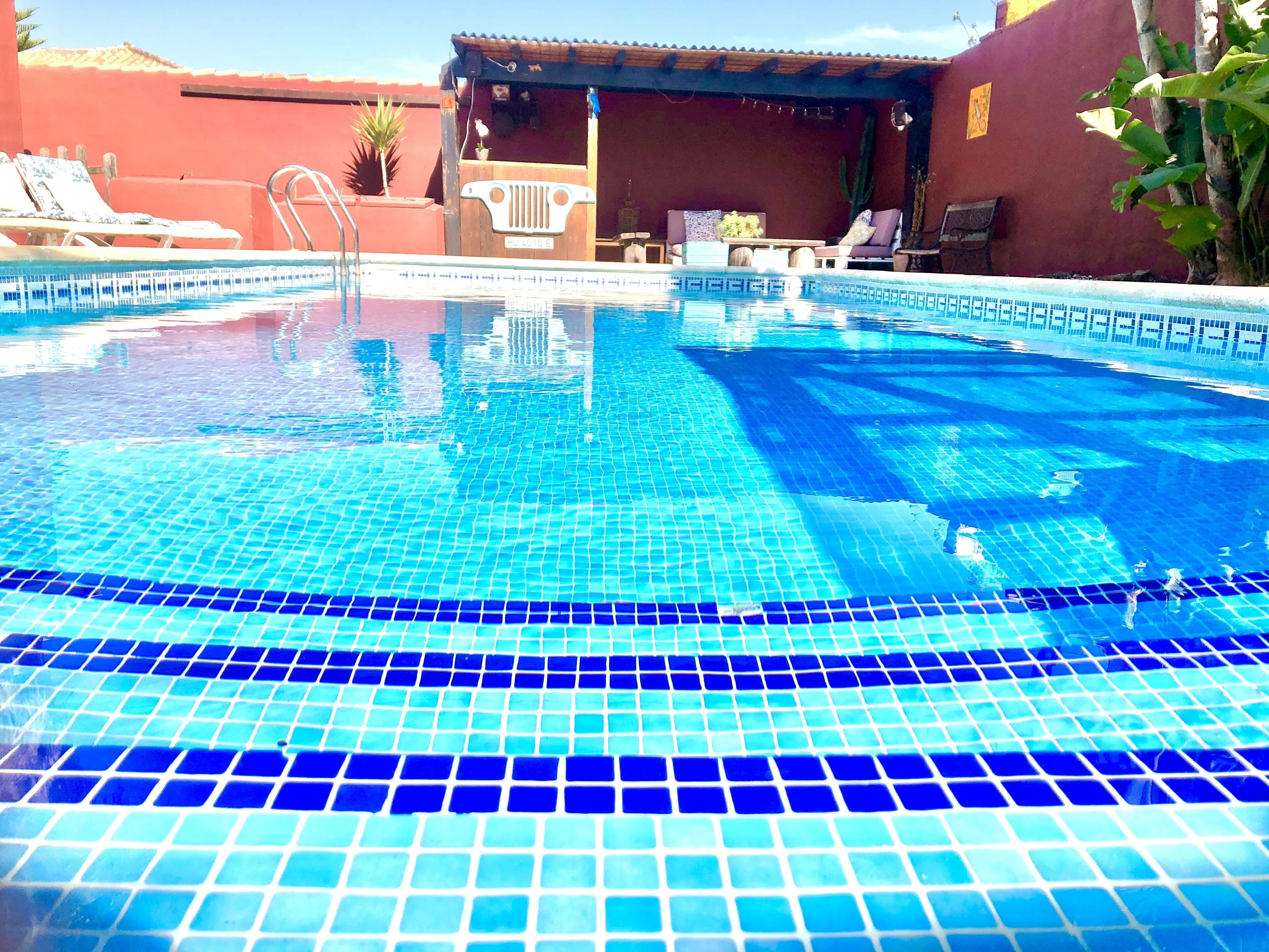 Ein Swimmingpool mit blauen Fliesen, einer Holzterrasse, Liegestühlen und einem überdachten Sitzbereich.