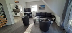 Un salon moderne avec un canapé noir, deux fauteuils noirs et une table basse. On y trouve également un coin cuisine avec des meubles blancs et un téléviseur à écran plat fixé au-dessus du comptoir. Une grande fenêtre laisse entrer la lumière naturelle, et des œuvres d’art sont exposées sur les murs.