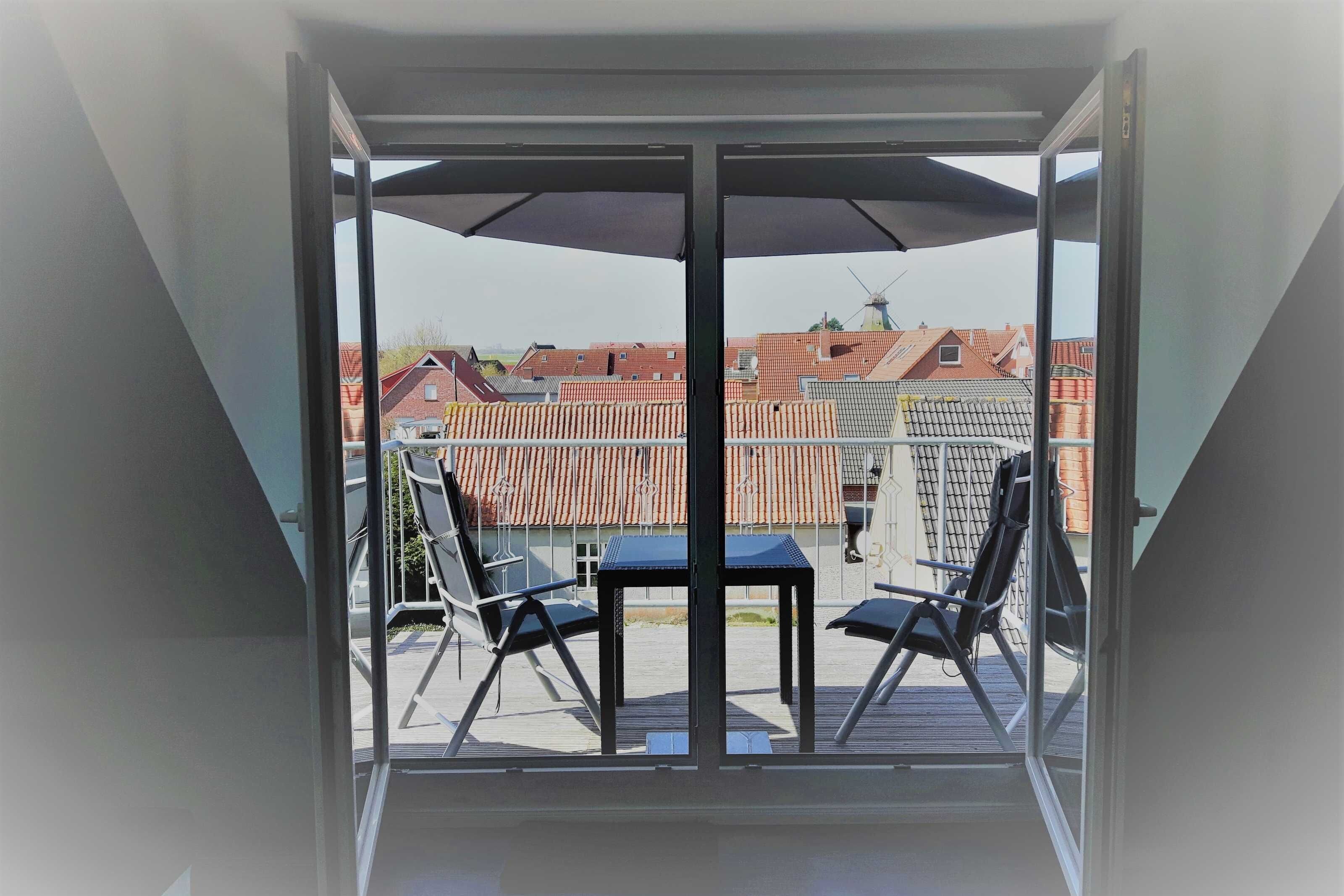 A balcony with a table and chairs, a large umbrella, and a view of a residential area with red-roofed buildings.