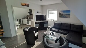 A modern living room with a black leather sofa, a glass coffee table, and a TV. There is a kitchen area with a dining table and chairs, and a window providing natural light. - Apartment Claudia (Wittmund)
