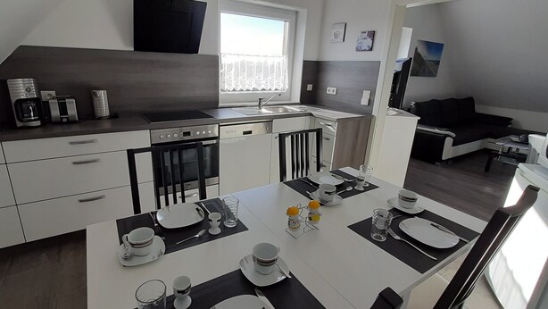 A modern kitchen with a dining area, featuring a built-in oven, a coffee machine, and a dining table set with plates, glasses, and cutlery. - Apartment Claudia (Wittmund)